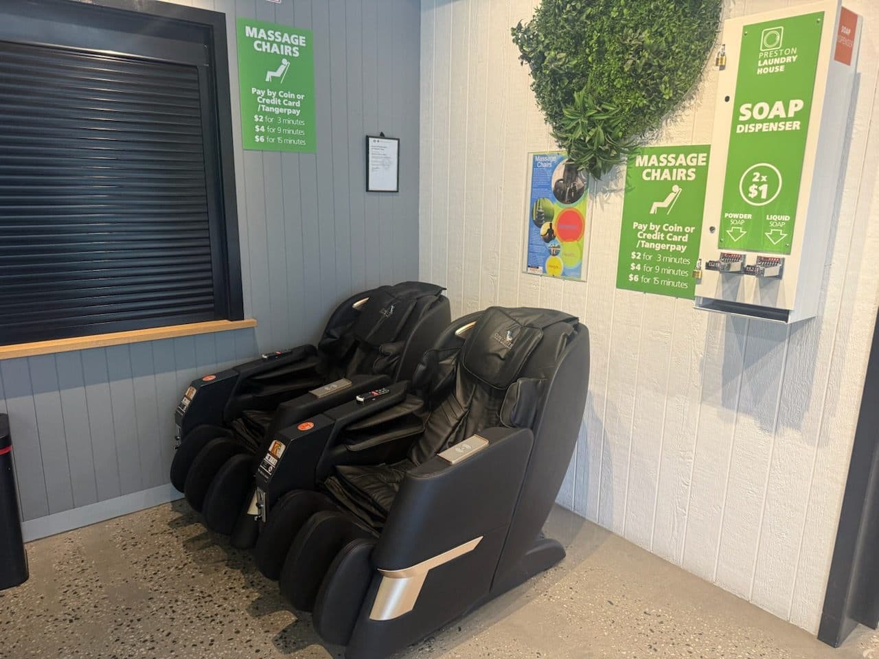 Two black reclining massage chairs against a light grey panelled wall at Preston Laundry House, beside a branded powder and liquid detergent dispenser and brand-green massage pricing signage